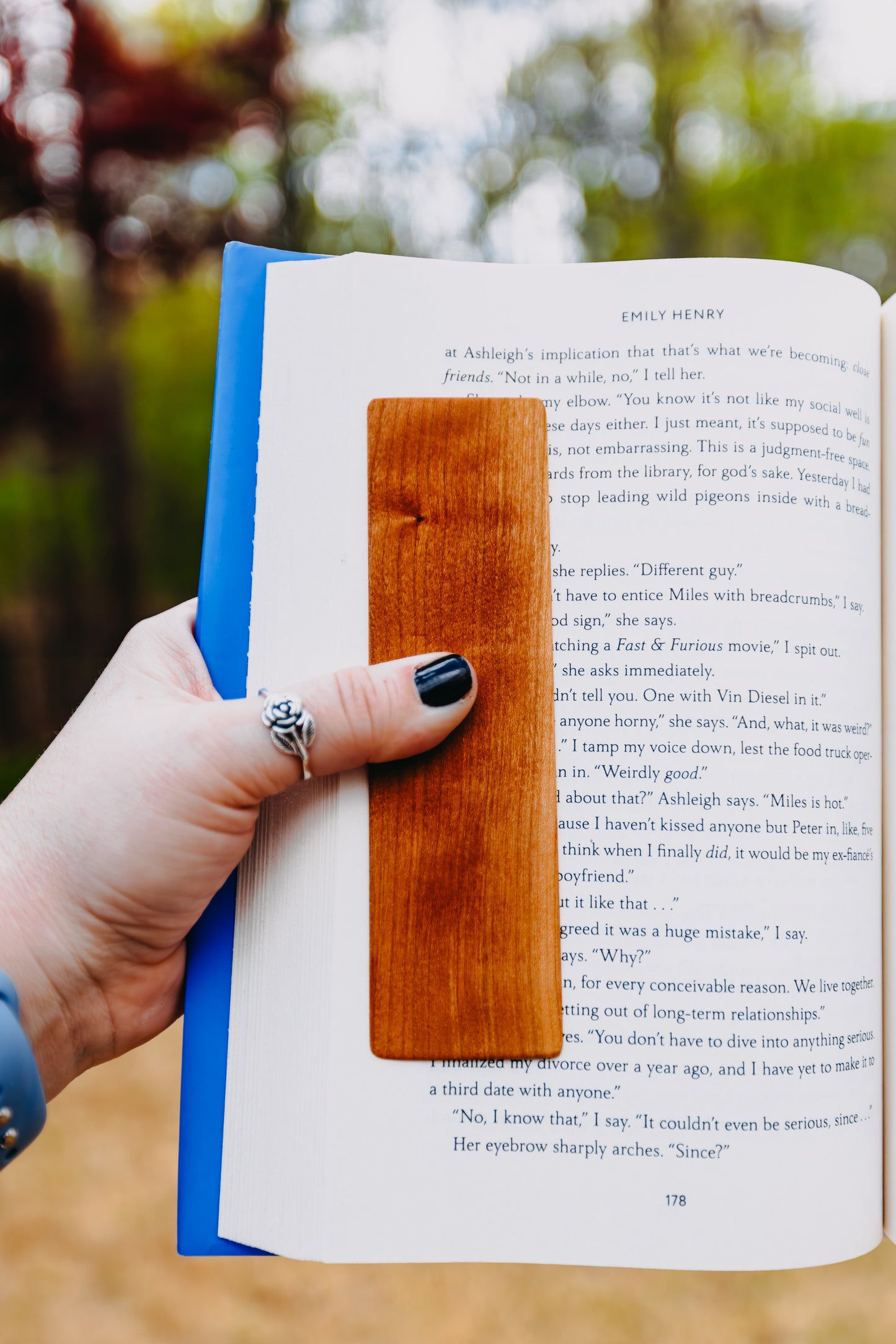 Hand holding an open book with a wooden cherry edge grain bookmark, outdoors.