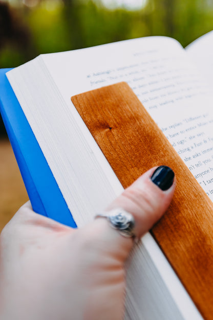 Hand holding a wooden cherry edge grain bookmark in an open book with a blurred green background