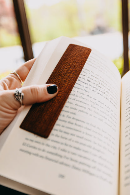 Hand holding an open book with a wooden sapele edge grain bookmark, blurred background